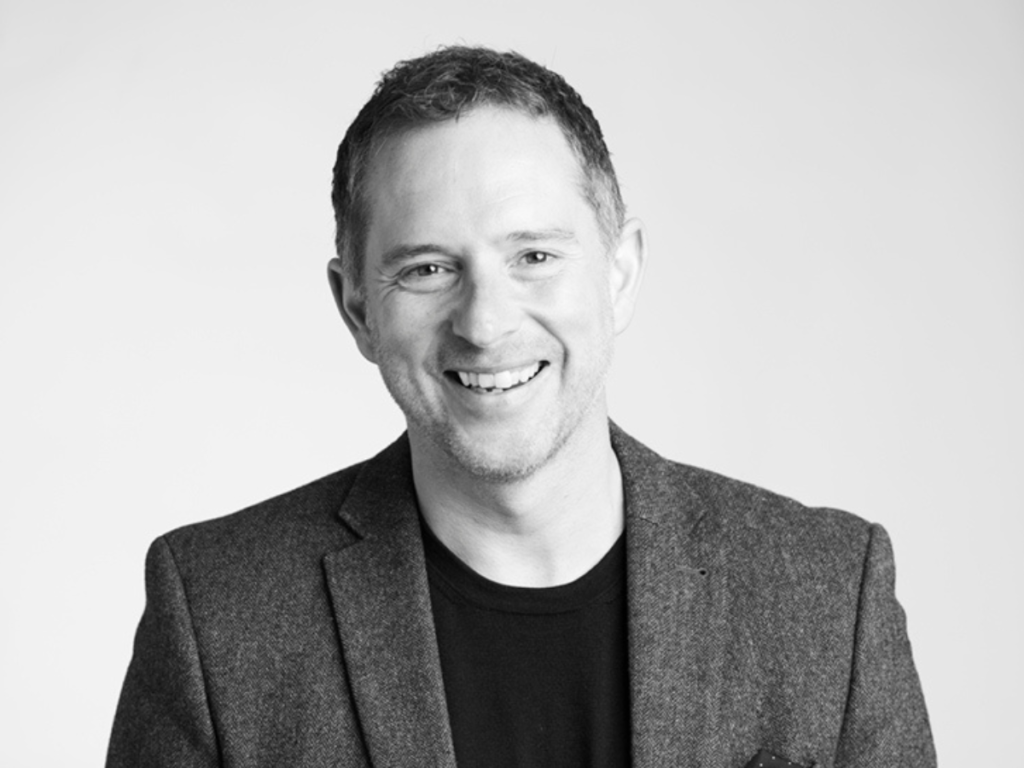 A black and white image of a man with short hair smiling. He's wearing a blazer over a black t-shirt 