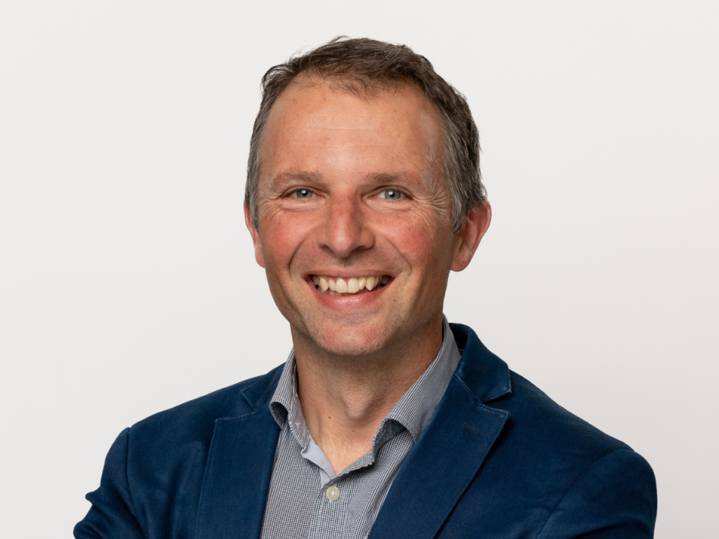 A man with short blonde hair smiles. He's wearing a navy blazer over a light blue button up shirt