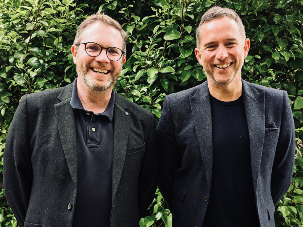 Two men in black suits smiling in front of a hedge