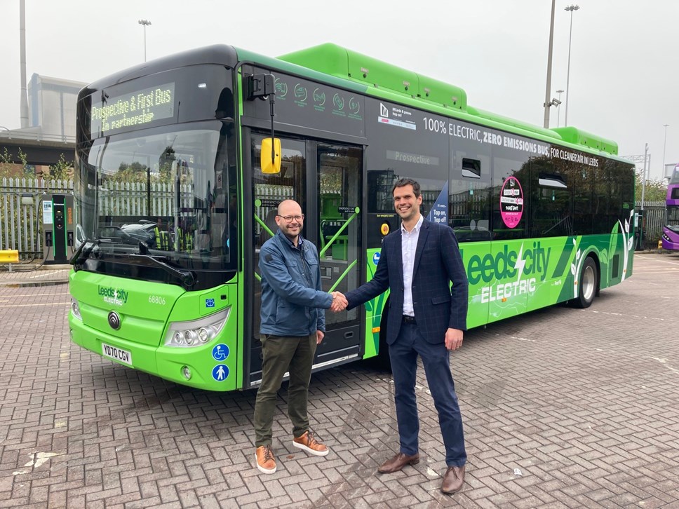 Two men shaking hands beside a green bus, with the destination sign reading 'Prospective & First Bus In Partnership'