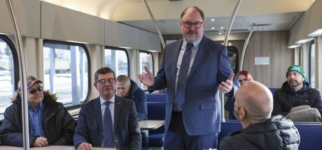 A businessman stands in the aisle of a train speaking to the passengers