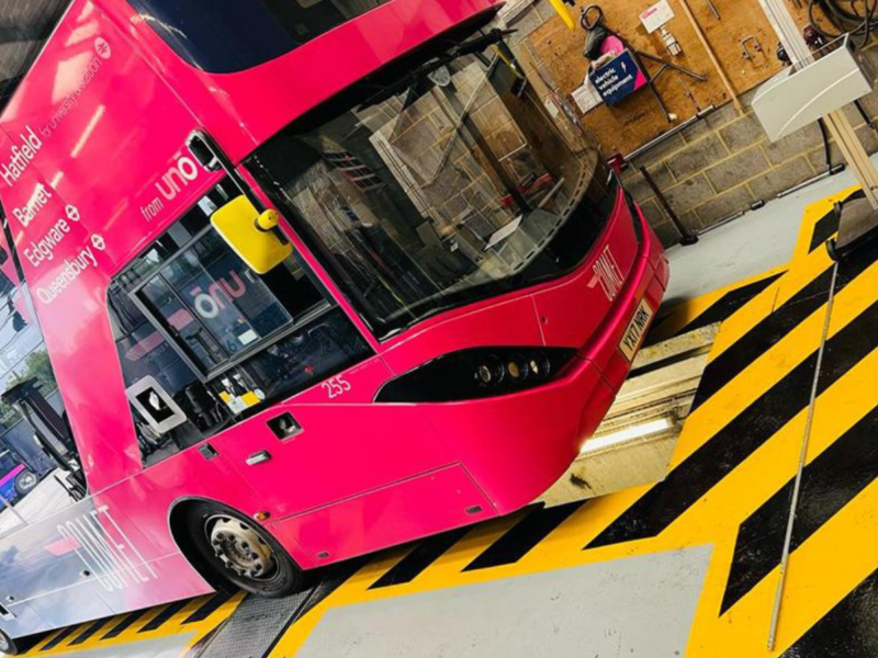 A bright pink double decker bus sits on top of a brake testing pit