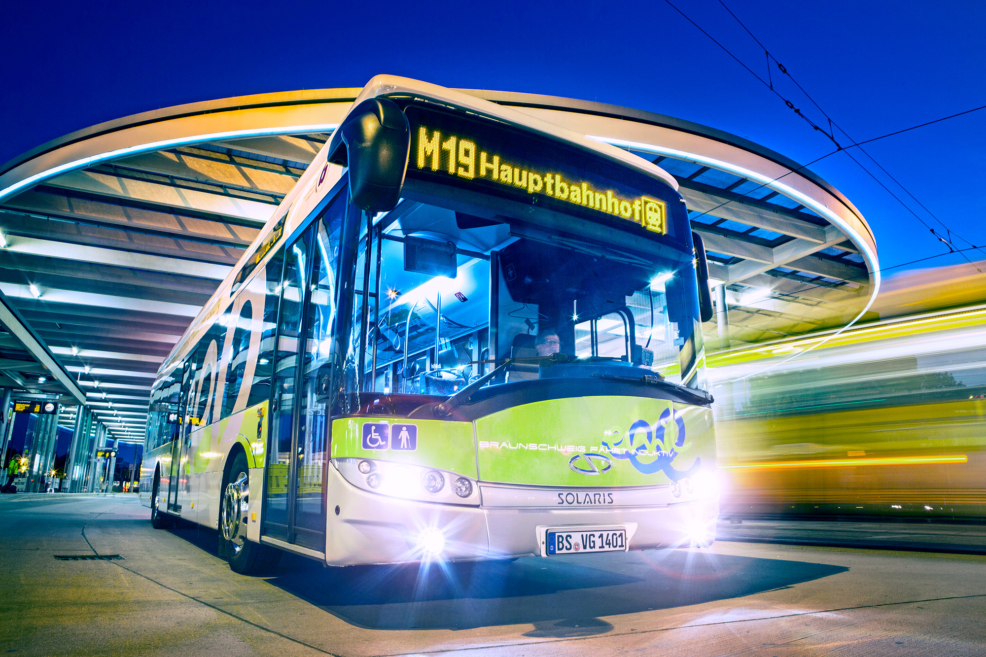 A green bus in motion at a train station, night time, with headlights on