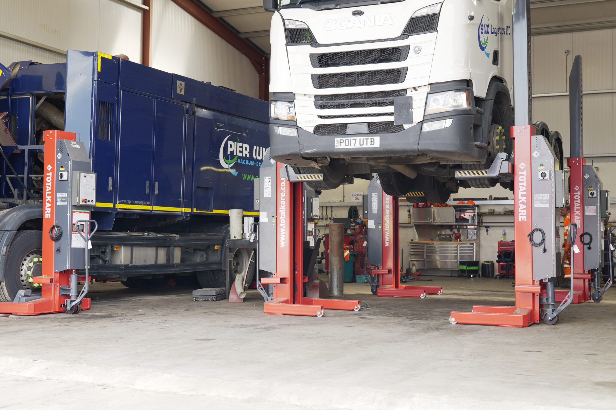 **Alt text:** A white Scania truck elevated on red Totalkare mobile column lifts inside a maintenance workshop, with a blue Pier UK vacuum excavation vehicle parked beside it and technicians working in the background.
