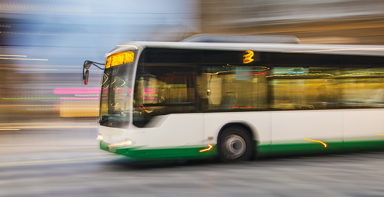 A white and green city bus in motion, captured with a blurred background to show speed, driving through an urban setting.