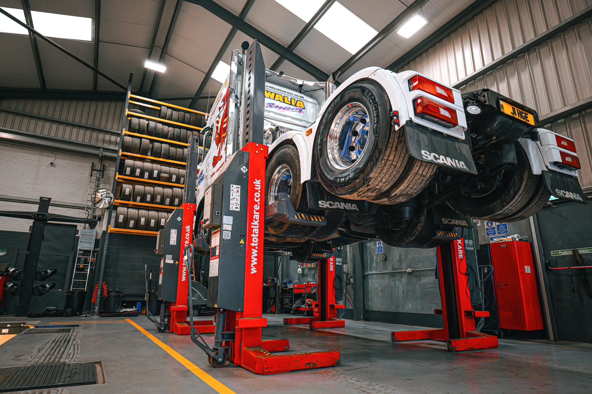 A large white Scania truck is elevated on bright red TotalKare vehicle lifts in a garage, with rows of tires on shelves in the background and a concrete floor with yellow markings.
