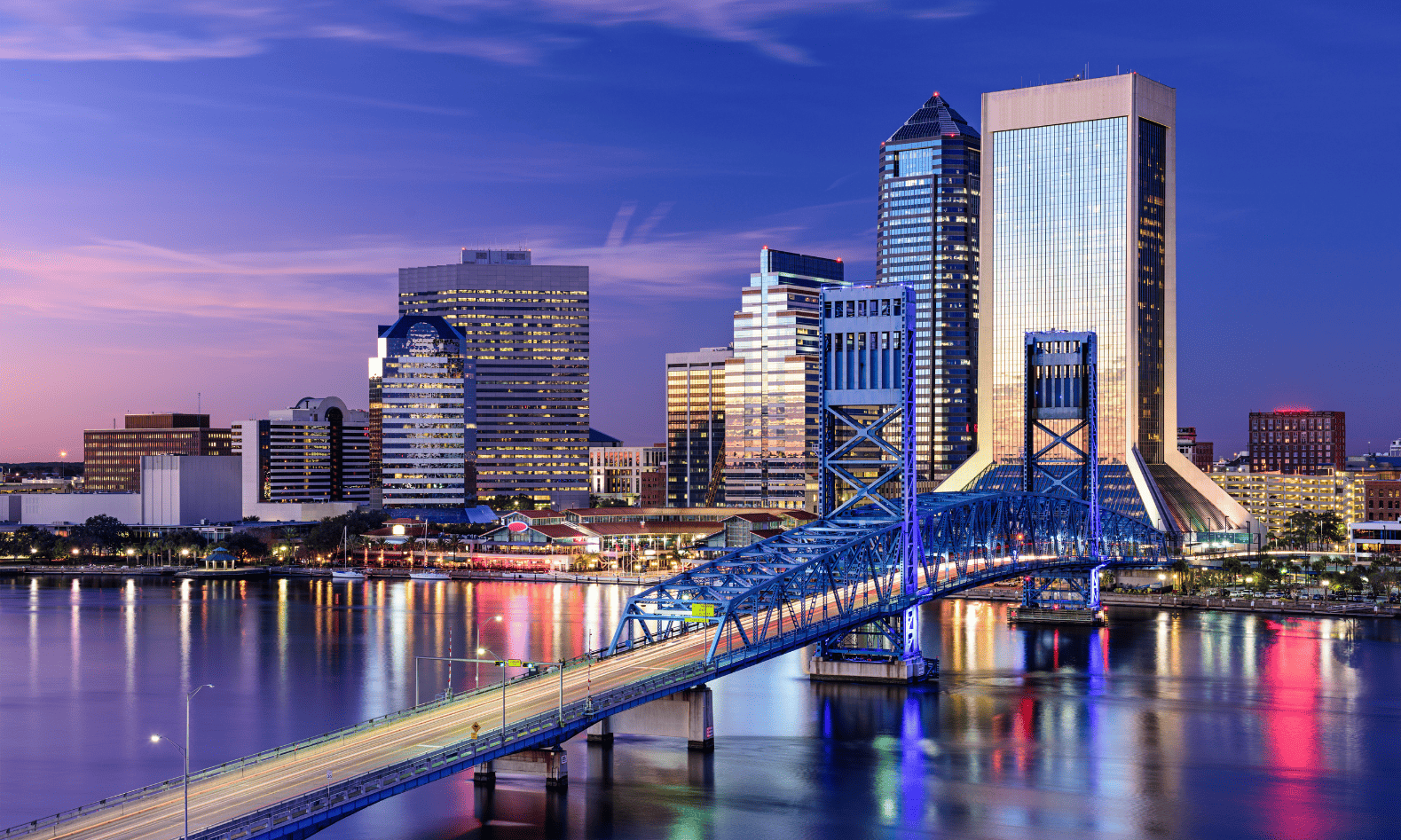 View of downtown Jacksonville, Florida at twilight, featuring illuminated skyscrapers and the Main Street Bridge reflecting on the St. Johns River.