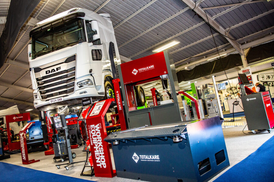 A bright workshop or exhibition hall features a large white DAF truck elevated on red vehicle lifts, surrounded by other industrial workshop equipment, including a blue box with "TOTALKARE" branding.
