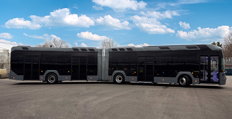 A long, modern grey articulated bus with dark tinted windows is parked outdoors on an asphalt surface under a blue sky with white clouds.