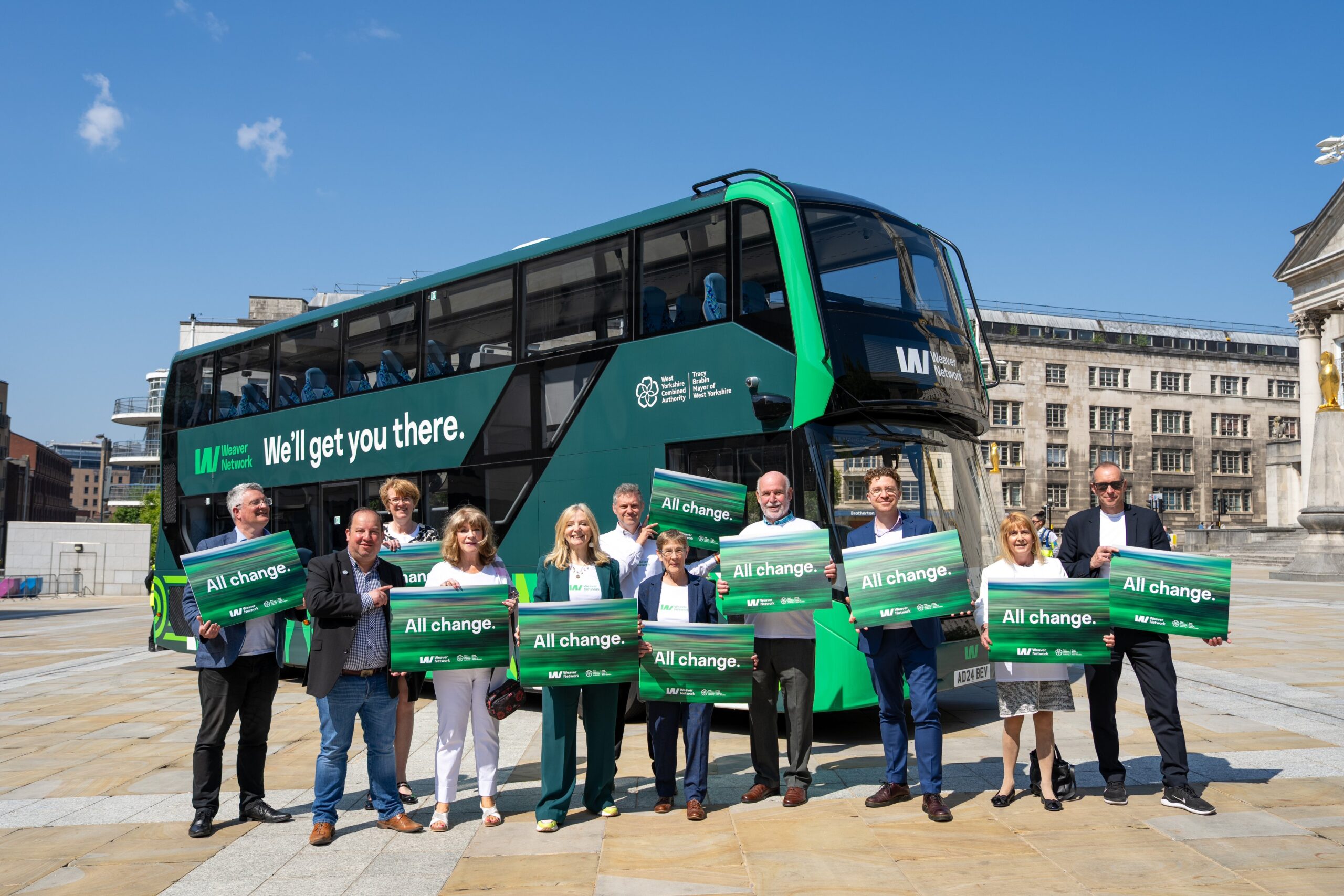 11 people stood in front of a green bus. They are smiling and hold signs that say All Change