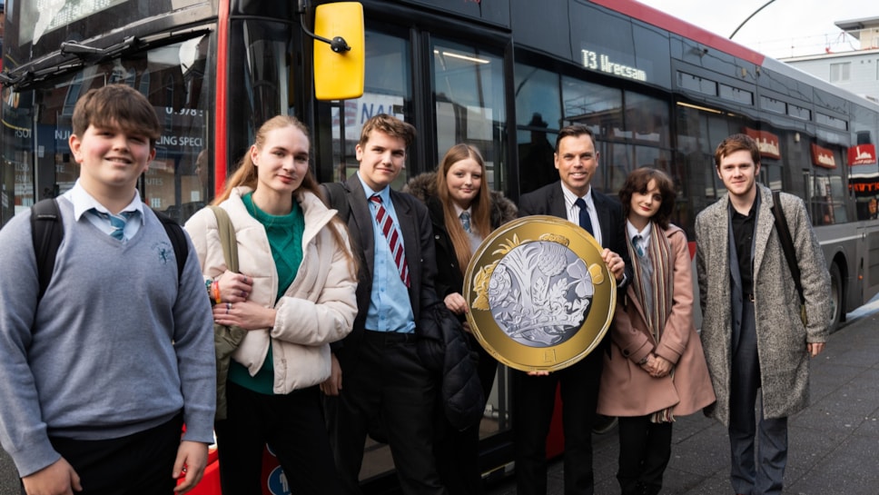 A group of people stood by a bus, one holding a massive £1 piece