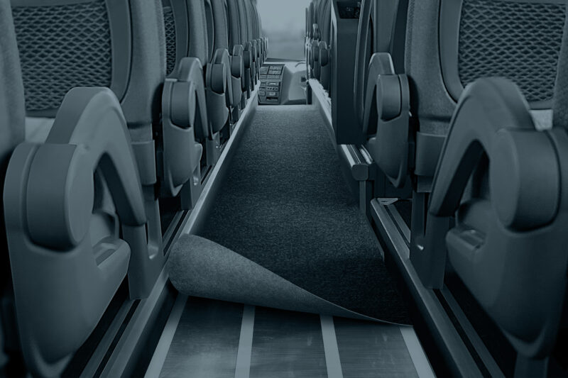 View down a coach aisle between rows of seats, with the grey carpet partly peeled back to reveal the metal floor and fastening strips underneath.