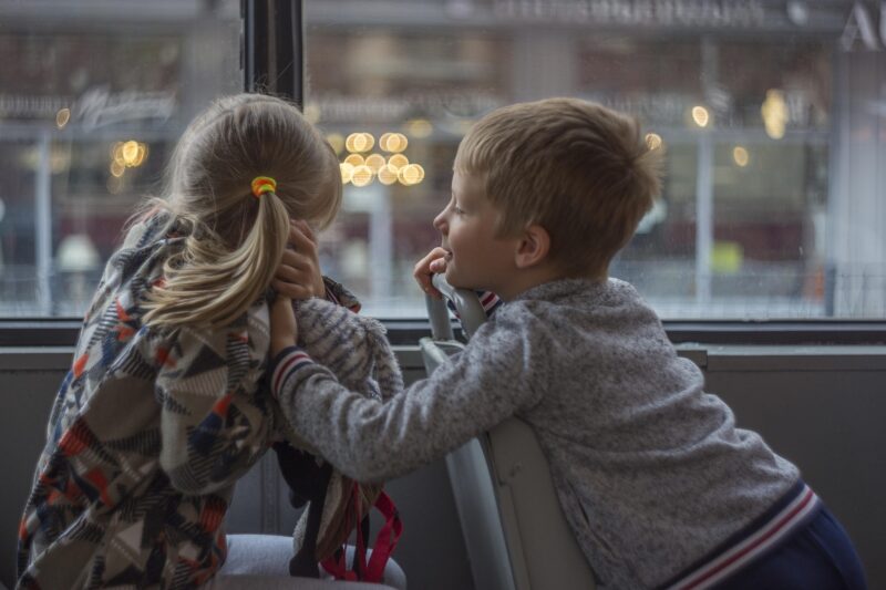 Two young children sitting and leaning on bus seats look out of a rain-streaked window at a city street, a reminder of the passengers who depend on emergency exits remaining usable when a bus is under strain
