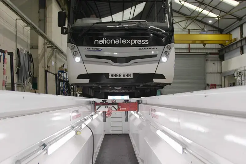 A National Express coach parked over a white-lined vehicle inspection pit in a workshop, seen from inside the pit. Strip lights run along both pit walls, a red pit jack spans the pit beneath the coach, and steps lead up out of the far end.