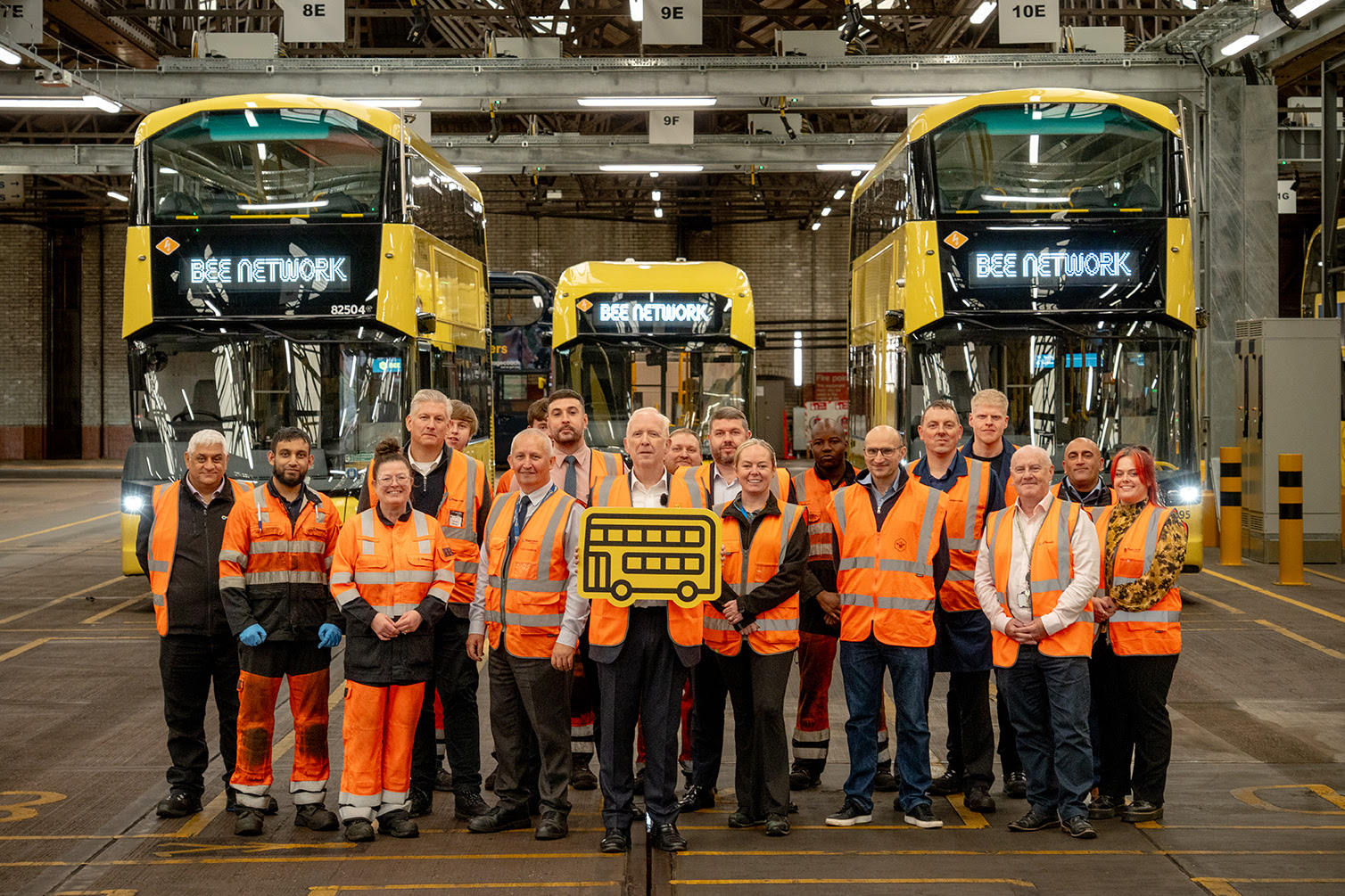 A group of people in orange high vis surrounding buses in a depot